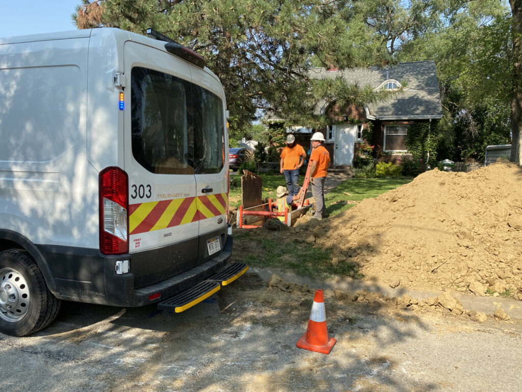 Two workers stand next to a hole where a third work is in the ground, on a ladder. Next to them is a large pile of dirt. They are in front of a home.