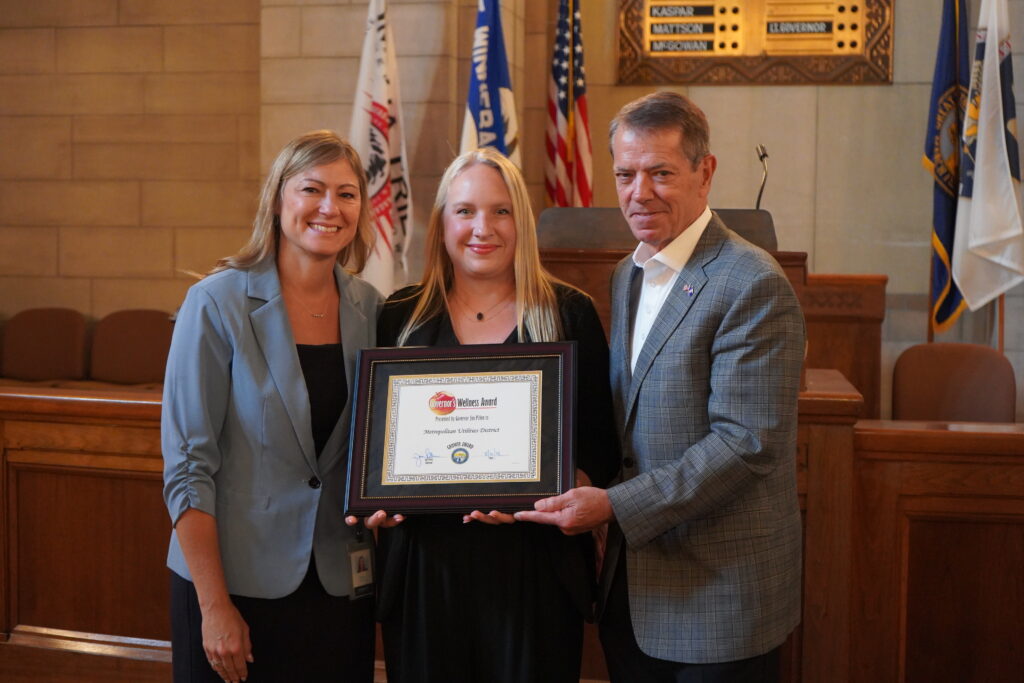 Two female M.U.D. employees smile while being handed award from Nebraska's governor in legislative chamber.