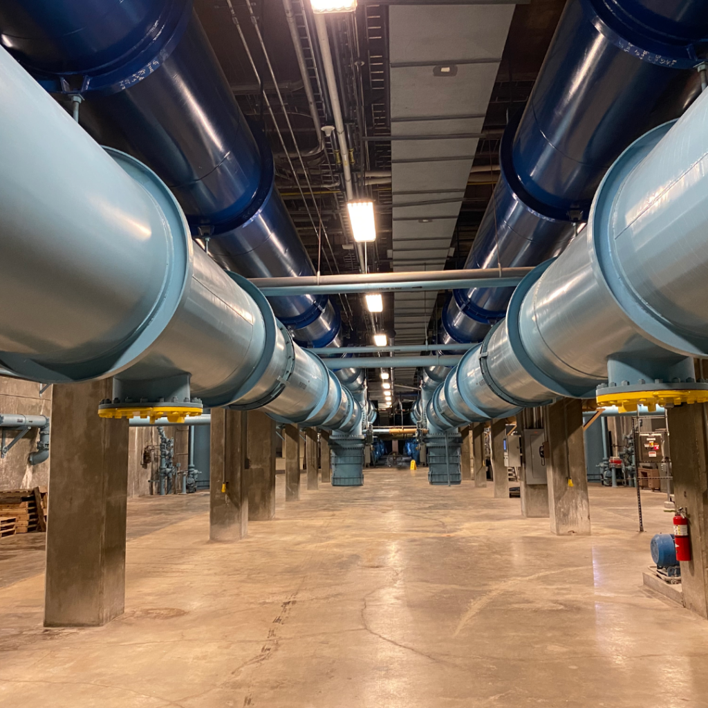Large water transmission pipes run through an underground utility facility with concrete supports and industrial equipment.